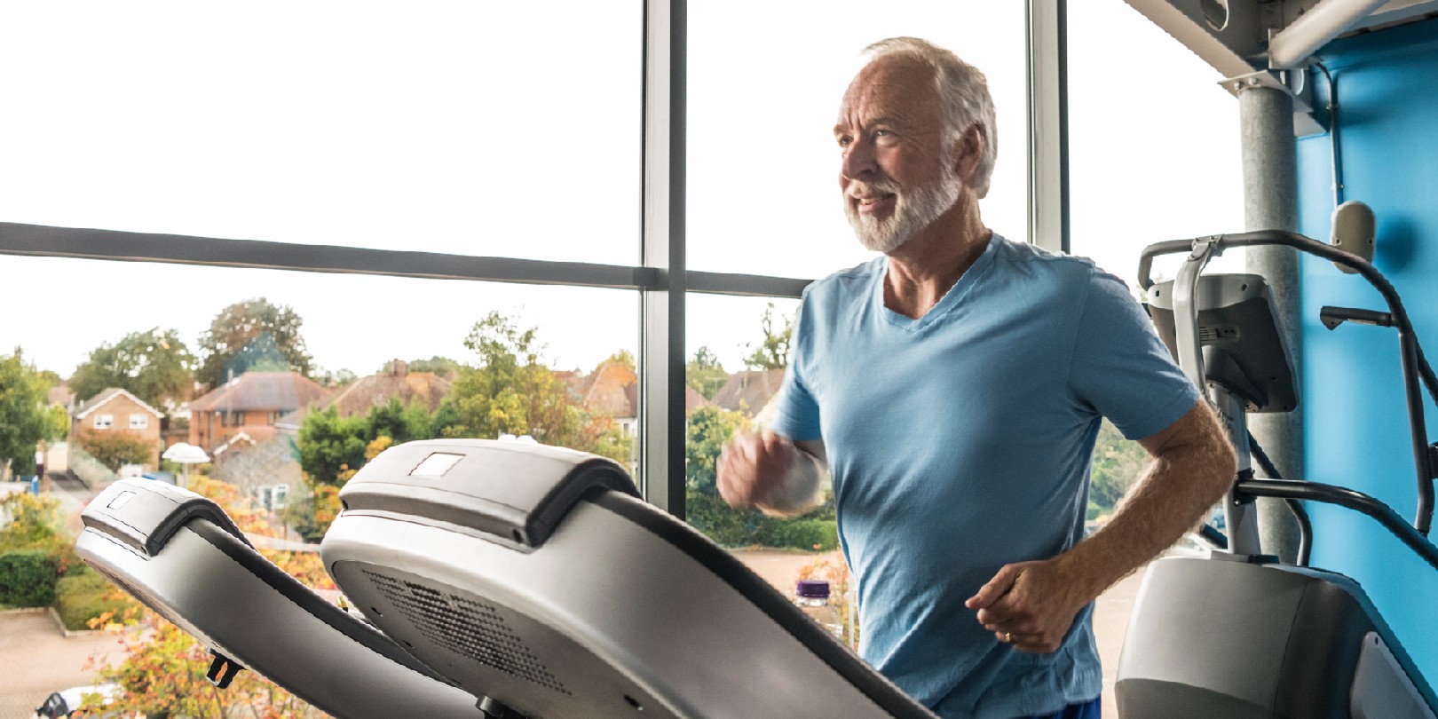 Man in his 60s running in the gym by a window, he is focused and healthy, with white hair and beard