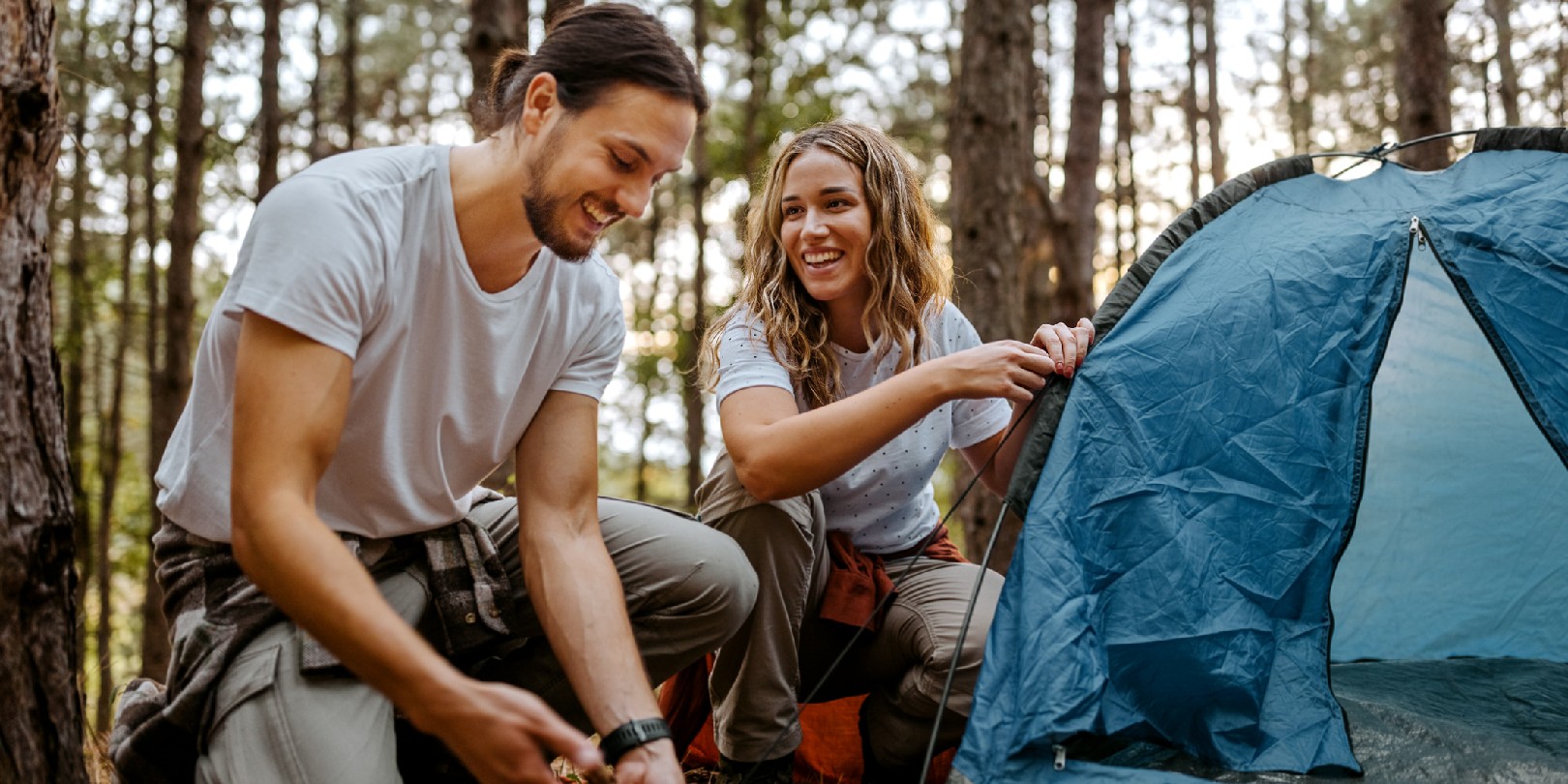 Beautiful smiling women with handsome boyfriend setting up tent in forest while talking