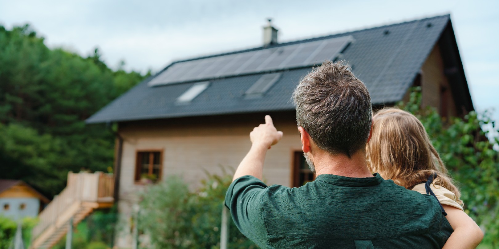 Rear view of dad holding her little girl in arms and showing at their house with solar panels.Alternative energy, saving resources and sustainable lifestyle concept.